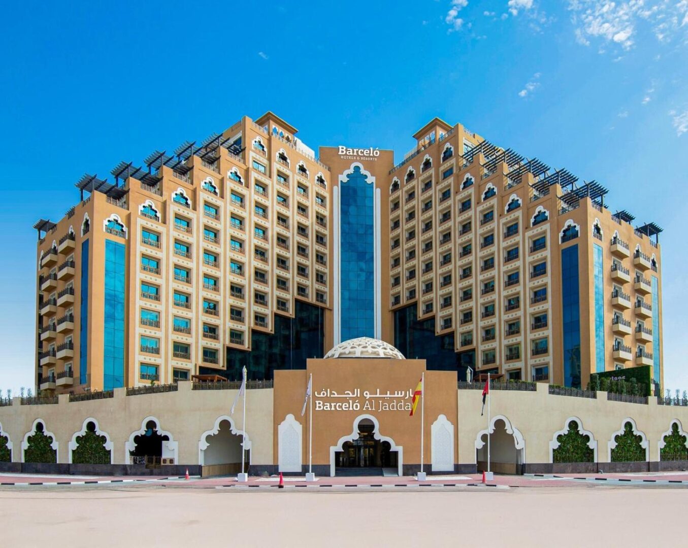 Daytime view of a contemporary hotel with a beige and white facade in Dubai. The building features a clean, geometric design with large windows and rooftop terraces. Palm trees and landscaped greenery are present in the foreground, creating a welcoming atmosphere