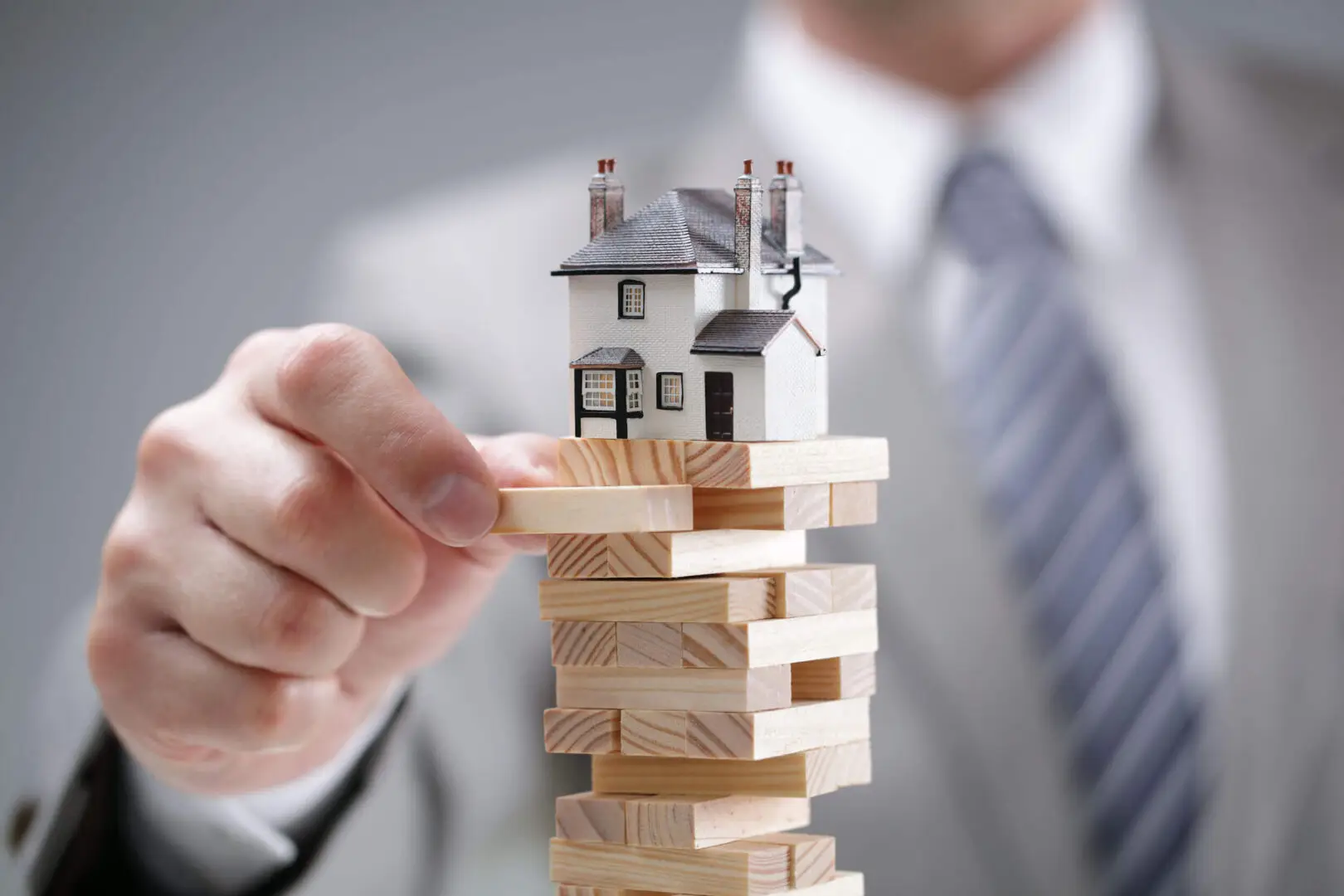 The image symbolizes the careful planning and strategy involved in real estate or construction, as a model house is placed atop a precarious stack of wooden blocks, similar to a Jenga game. This visual emphasizes the balance needed in property investment or development.