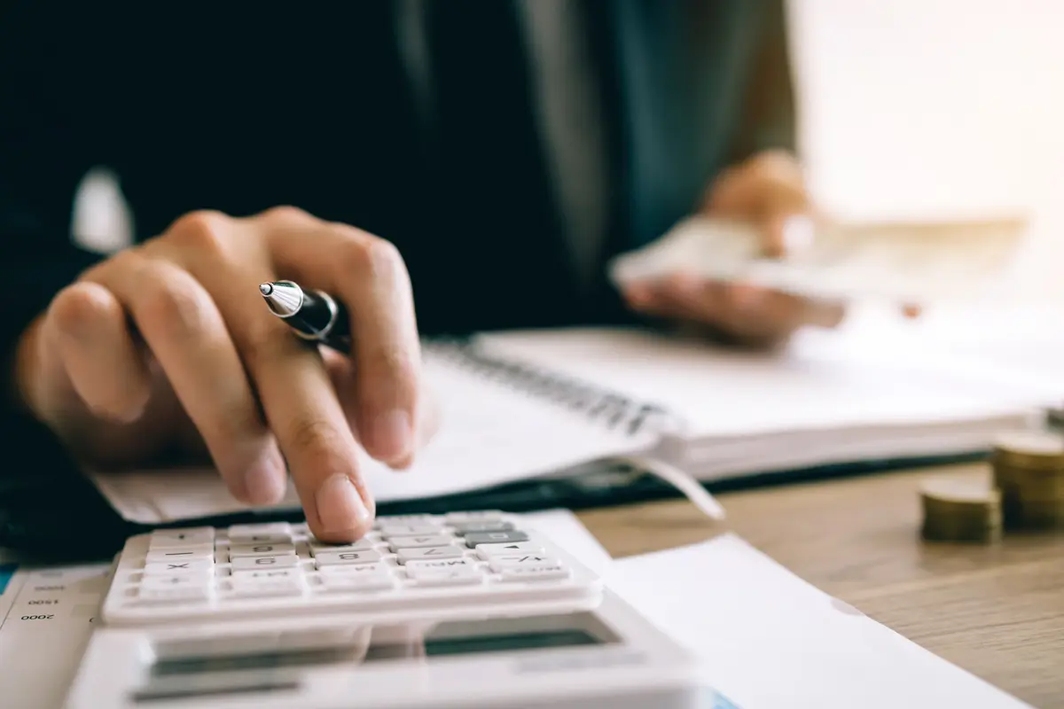 An image of a person writing on a piece of paper with a pen in their hand, with a calculator in front of them, working