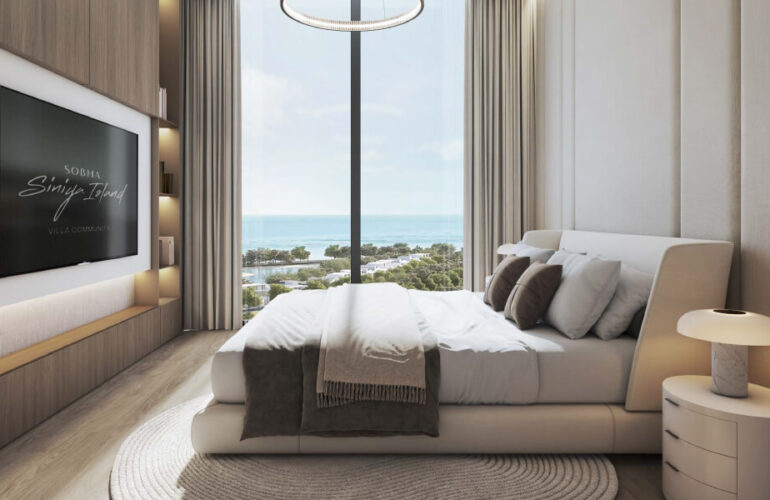 A bedroom in Pierside Marina Residences with a double bed. Next to the bed, there's a large floor-to-ceiling window with a view of the sea and beach. There is also a TV facing the bed.